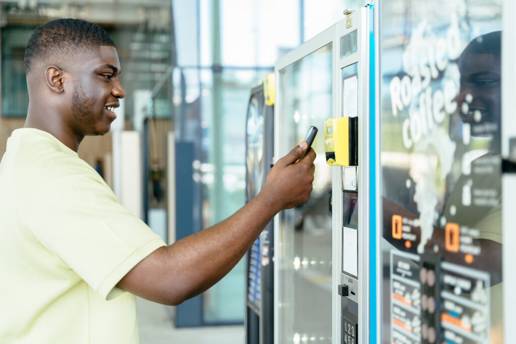 Paying With Mobile Phone. Young Traveler Black Man Choosing A Snack Or Drink At Vending Machine